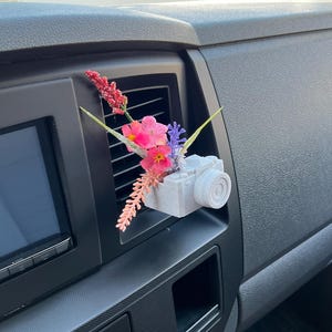 May include: A gray camera-shaped car air freshener with a bouquet of pink, red, and purple flowers. The air freshener is attached to a car vent, adding a decorative touch to the vehicle's interior.