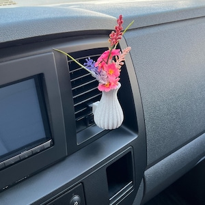 May include: A small, white, textured vase filled with colourful artificial flowers, including pink, red, and purple blooms. The vase is designed to clip onto a car's air vent, adding a decorative touch to the vehicle's interior.