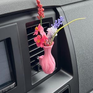 May include: A pink, 3D-printed car vent vase holding a small bouquet of colorful artificial flowers. The flowers include pink, purple, and red blooms. The vase is attached to a black car vent.