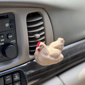 May include: A tan-colored chicken air vent clip, with a red comb and wattle, is attached to a car's air vent. The chicken is positioned in front of the vent, adding a decorative touch to the vehicle's interior.