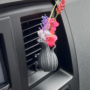 May include: A grey, tulip-shaped car vent clip holding colourful artificial flowers. The flowers include pink, red, purple, and white blooms. The clip is inserted into a black car vent.