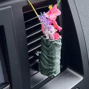May include: A dark green, cylindrical car air vent diffuser with a wavy texture, holding colourful artificial flowers. The flowers include pink, purple, and red blossoms, adding a touch of nature to the car's interior.