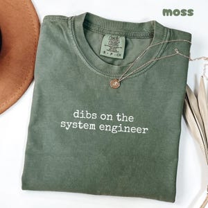 May include: A moss green t-shirt with the phrase "dibs on the system engineer" printed in white. The shirt has a crew neck and short sleeves. A gold necklace with a pendant is draped on the shirt.