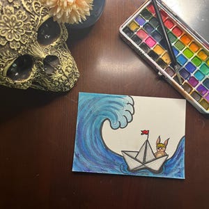 May include: A watercolour painting of a paper boat sailing on a blue wave, with a rabbit wearing a construction hat. A skull, flowers in a blue vase, and a watercolour paint set are in the background.
