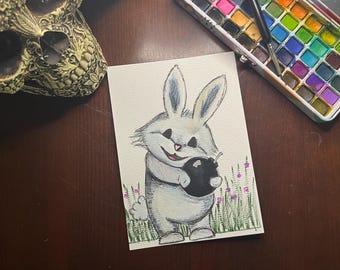 May include: Watercolor painting of a gray bunny holding a black object, set against a backdrop of green grass and purple flowers. The artwork is on a white rectangular card. A metal watercolor paint set and a decorative skull are also visible.