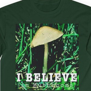 May include: Dark green long-sleeve shirt with a square graphic featuring a mushroom in green grass. The graphic includes the text "I BELIEVE (in Mushrooms)" in white. The shirt is made of a soft, comfortable material.