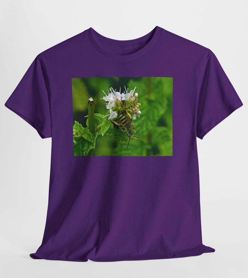 May include: A purple t-shirt featuring a nature-themed graphic. The graphic is a close-up of a green grasshopper on a white flower with green leaves in the background. The t-shirt is made of a soft material.