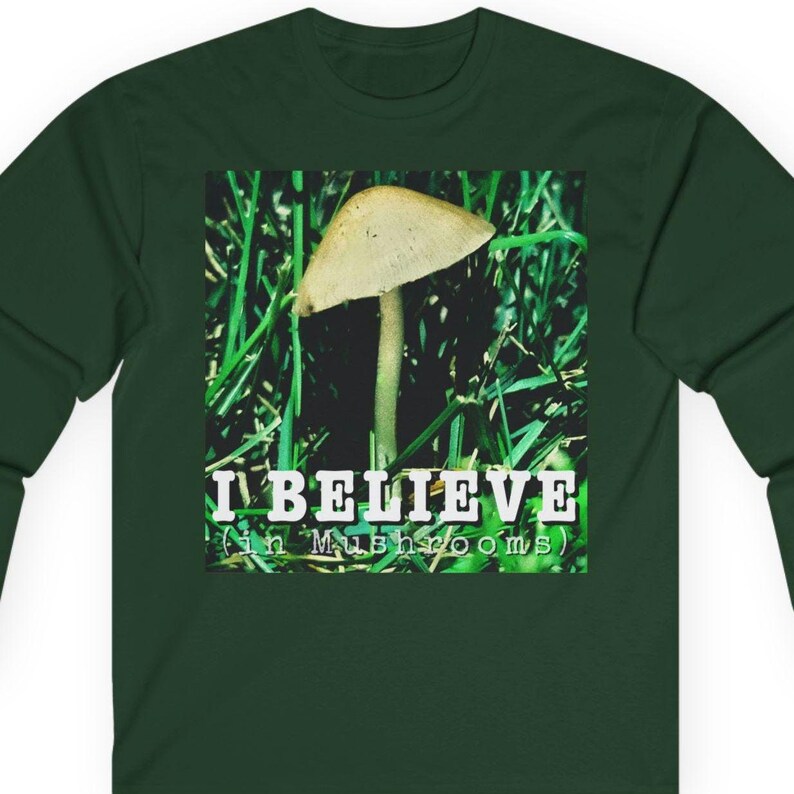 May include: Dark green long-sleeve shirt with a square graphic featuring a mushroom in green grass. The graphic includes the text "I BELIEVE (in Mushrooms)" in white. The shirt is made of a soft, comfortable material.