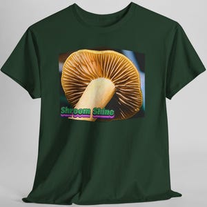 May include: A dark green short-sleeved t-shirt featuring a central graphic of a mushroom with visible gills. The text "Shroom Shine" is printed in green and purple lettering at the bottom of the mushroom illustration on the front of the shirt.