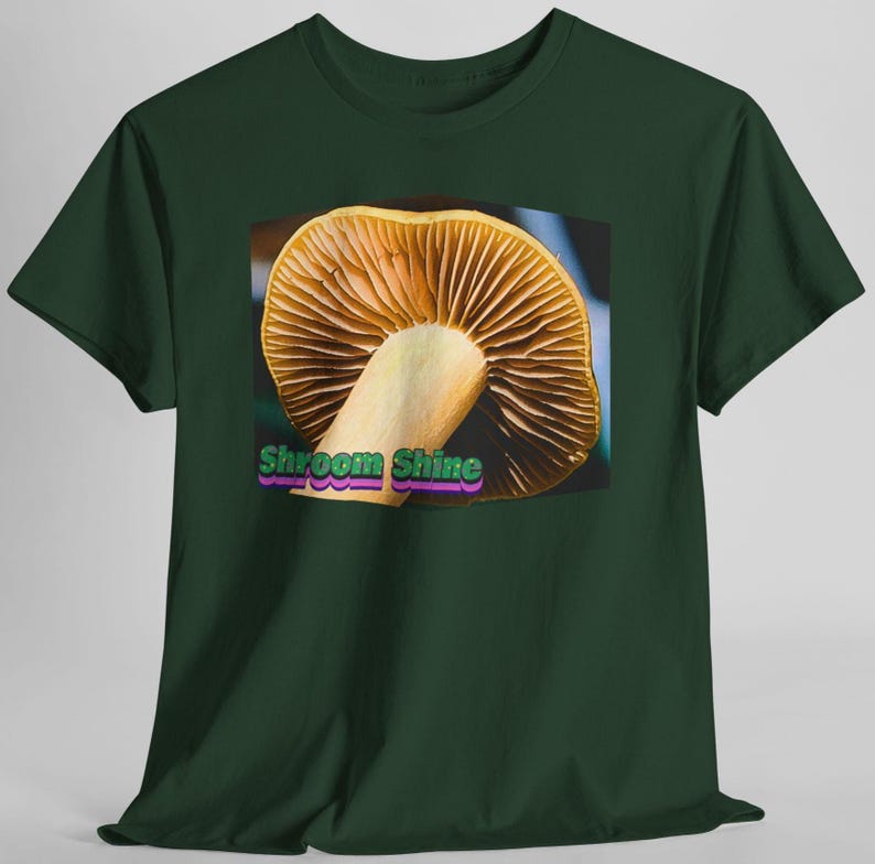 May include: A dark green short-sleeved t-shirt featuring a central graphic of a mushroom with visible gills. The text "Shroom Shine" is printed in green and purple lettering at the bottom of the mushroom illustration on the front of the shirt.