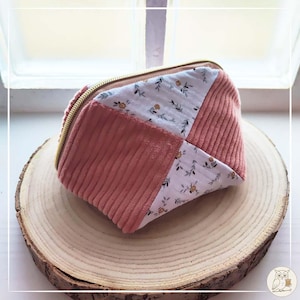 Peut inclure: Une petite trousse de maquillage matelassée avec une fermeture éclair dorée. Le sac présente un motif patchwork avec des sections de tissu rose côtelé et de tissu blanc à motifs floraux. Le sac est posé sur une tranche de bois.