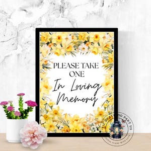 May include: A framed sign with the text "PLEASE TAKE ONE In Loving Memory" surrounded by yellow daffodils. A small white pot with pink flowers and a pink peony are in the foreground.
