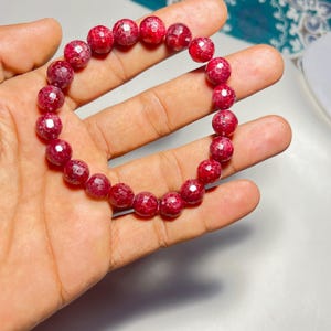Może przedstawiać: Czerwona bransoletka z kamieni szlachetnych z fasetowanymi, okrągłymi koralikami. Koraliki mają głęboki, bogaty czerwony kolor, a bransoletka jest nawleczona na elastyczną opaskę. Bransoletka jest pokazana na dłoni.