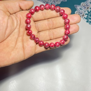 Może przedstawiać: Czerwona bransoletka z koralików z nakrapianym wzorem. Bransoletka wykonana jest z okrągłych koralików i trzymana jest w dłoni. Koraliki mają średnicę około 0,8 cm. To biżuteria.