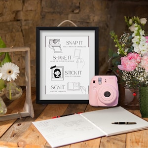 May include: A pink instant camera sits on a wooden table next to a framed sign with instructions: "Snap it, Shake it, Stick it, Sign it." A notebook, pen, and flowers complete the scene. A wooden frame holds flowers in glass vases.
