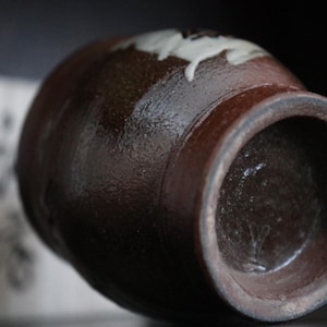 May include: A brown ceramic jar with a wide opening and a rounded body. The jar has a dark brown exterior with a lighter brown interior. A white floral design is visible near the top. The jar is likely handmade and has a rustic aesthetic.