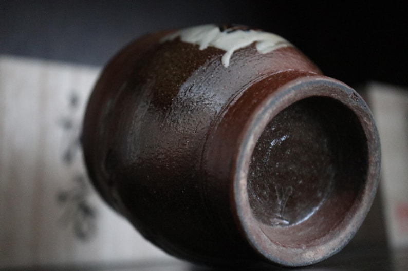 May include: A brown ceramic jar with a wide opening and a rounded body. The jar has a dark brown exterior with a lighter brown interior. A white floral design is visible near the top. The jar is likely handmade and has a rustic aesthetic.