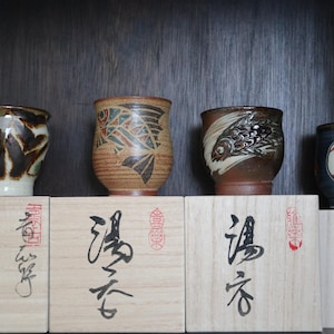 May include: Four ceramic teacups with distinctive designs, including abstract patterns and fish motifs. The cups are displayed on wooden boxes with Japanese calligraphy. The teacups vary in colour, with brown, blue, and white accents.