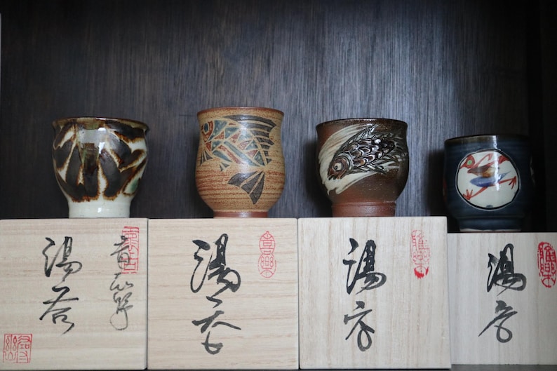 May include: Four ceramic teacups with distinctive designs, including abstract patterns and fish motifs. The cups are displayed on wooden boxes with Japanese calligraphy. The teacups vary in colour, with brown, blue, and white accents.