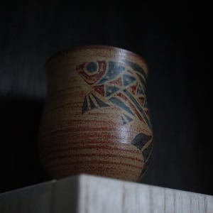 May include: A handmade ceramic cup with a fish design. The cup has a beige base with red and blue geometric patterns. The cup is cylindrical and sits on a wooden surface.