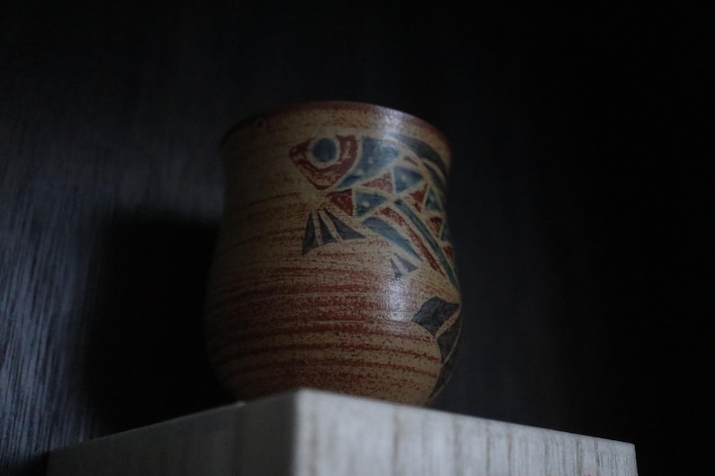 May include: A handmade ceramic cup with a fish design. The cup has a beige base with red and blue geometric patterns. The cup is cylindrical and sits on a wooden surface.