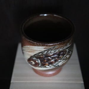 May include: A handcrafted ceramic cup with a fish design. The cup features a brown and white colour scheme with a detailed fish motif on the side. The cup is approximately 7.6 cm tall and 7.6 cm in diameter, with a dark interior.