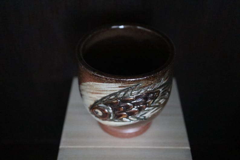 May include: A handcrafted ceramic cup with a fish design. The cup features a brown and white colour scheme with a detailed fish motif on the side. The cup is approximately 7.6 cm tall and 7.6 cm in diameter, with a dark interior.