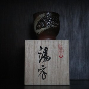 May include: A brown and white ceramic cup with a fish design rests on a wooden box with Japanese calligraphy. The cup's design is detailed, and the box has a red stamp. The cup appears to be handmade, with a unique shape.