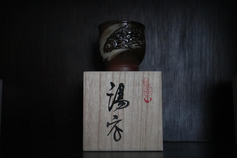 May include: A brown and white ceramic cup with a fish design rests on a wooden box with Japanese calligraphy. The cup's design is detailed, and the box has a red stamp. The cup appears to be handmade, with a unique shape.