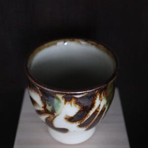 May include: A ceramic teacup with a white base and a brown, green, and white patterned exterior. The rim is dark brown. The cup is set against a dark background.