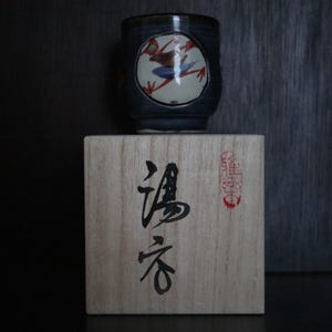 May include: A dark blue ceramic teacup with a frog design in a white circle, resting on a wooden box with Japanese characters. The frog is red, white, and blue. The box has Japanese text and a red stamp.
