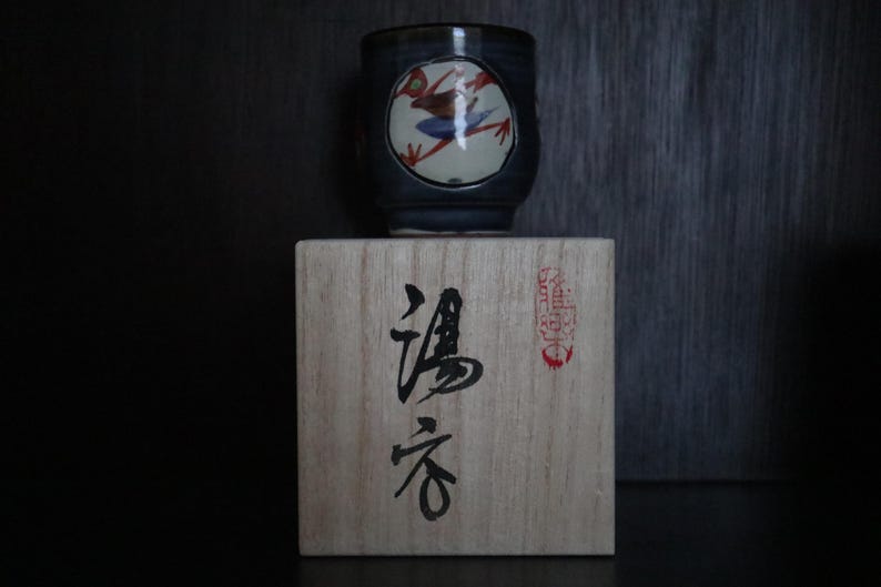 May include: A dark blue ceramic teacup with a frog design in a white circle, resting on a wooden box with Japanese characters. The frog is red, white, and blue. The box has Japanese text and a red stamp.