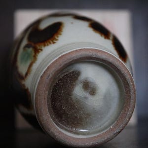 May include: A ceramic vase with a rounded body and a textured, unglazed base. The vase has a cream-coloured background with brown and green abstract patterns. The rim and base have a dark brown, earthy tone, suggesting a handcrafted aesthetic.