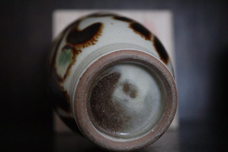 May include: A ceramic vase with a rounded body and a textured, unglazed base. The vase has a cream-coloured background with brown and green abstract patterns. The rim and base have a dark brown, earthy tone, suggesting a handcrafted aesthetic.