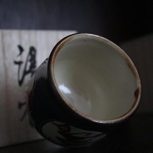 May include: A ceramic teacup with a dark blue exterior and a light interior. The rim is brown. The cup has a floral design. A wooden box with Japanese characters is in the background.