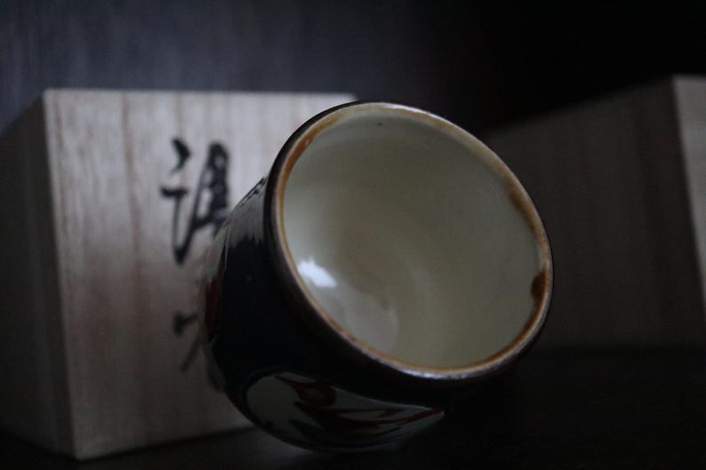 May include: A ceramic teacup with a dark blue exterior and a light interior. The rim is brown. The cup has a floral design. A wooden box with Japanese characters is in the background.