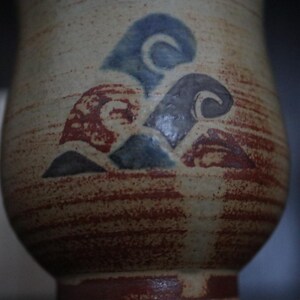 May include: A ceramic vase with a textured, beige surface and a reddish-brown base. The vase features a design of blue, grey, and red abstract shapes. The design is reminiscent of waves or clouds. The vase has a rounded shape.