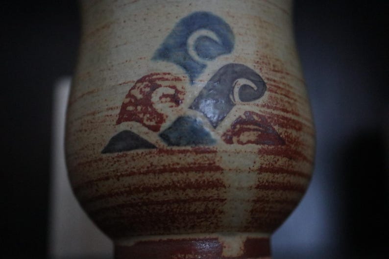May include: A ceramic vase with a textured, beige surface and a reddish-brown base. The vase features a design of blue, grey, and red abstract shapes. The design is reminiscent of waves or clouds. The vase has a rounded shape.