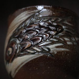 May include: A handcrafted ceramic mug with a dark brown glaze and a detailed relief design. The design features a fish motif with a glossy finish. The mug has a unique, textured appearance.