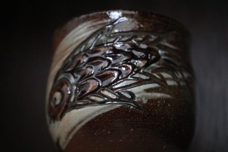 May include: A handcrafted ceramic mug with a dark brown glaze and a detailed relief design. The design features a fish motif with a glossy finish. The mug has a unique, textured appearance.