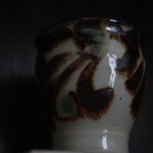 May include: A ceramic cup with a wide base and a slightly flared rim. The cup is off-white with brown and dark green abstract patterns. The cup is likely handmade and has a rustic aesthetic.