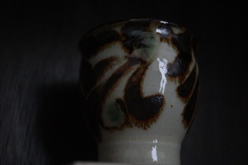 May include: A ceramic cup with a wide base and a slightly flared rim. The cup is off-white with brown and dark green abstract patterns. The cup is likely handmade and has a rustic aesthetic.