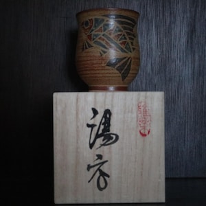 May include: A brown ceramic cup with a fish design in black, red, and green sits on a wooden block with Japanese characters. The cup has a textured surface and a dark brown rim. The wooden block has Japanese text and a red seal.