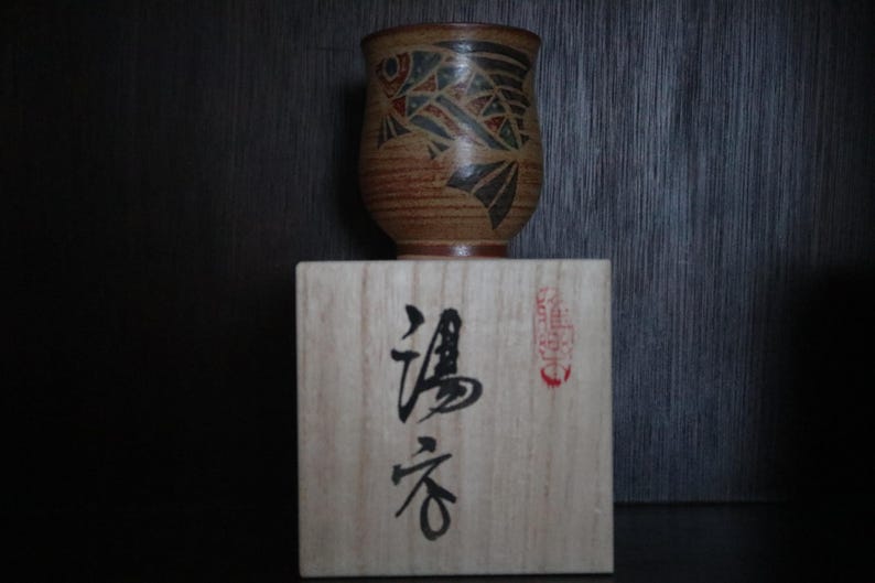 May include: A brown ceramic cup with a fish design in black, red, and green sits on a wooden block with Japanese characters. The cup has a textured surface and a dark brown rim. The wooden block has Japanese text and a red seal.