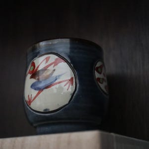 May include: A ceramic teacup with a dark blue glaze and a hand-painted design. The design features a bird with red, blue, and green accents on a white background. The cup is cylindrical and sits on a shelf.