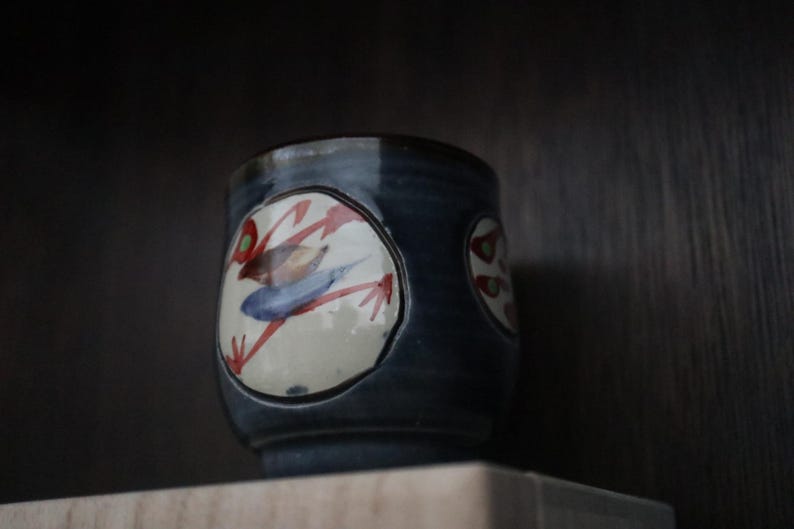 May include: A ceramic teacup with a dark blue glaze and a hand-painted design. The design features a bird with red, blue, and green accents on a white background. The cup is cylindrical and sits on a shelf.