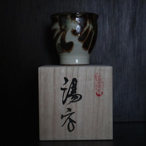 May include: A ceramic teacup with a brown and cream abstract design sits on a wooden box. The box features Japanese characters and a red stamp. The teacup is a traditional Japanese style.