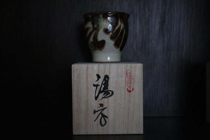 May include: A ceramic teacup with a brown and cream abstract design sits on a wooden box. The box features Japanese characters and a red stamp. The teacup is a traditional Japanese style.