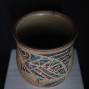 May include: A handcrafted ceramic mug with a fish design. The mug has a beige base with a fish illustration in red, blue, and black geometric shapes. The rim and interior are dark brown.