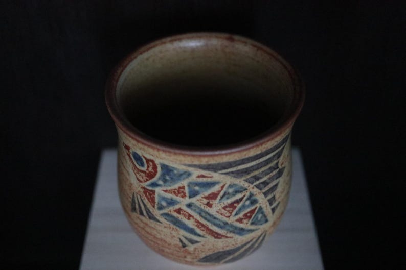 May include: A handcrafted ceramic mug with a fish design. The mug has a beige base with a fish illustration in red, blue, and black geometric shapes. The rim and interior are dark brown.